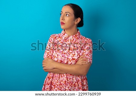 Charming thoughtful beautiful woman wearing floral dress over blue studio background stands with arms folded concentrated somewhere with pensive expression thinks what to do