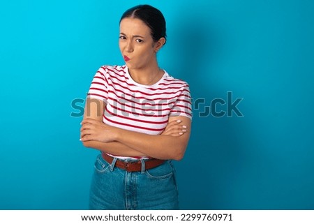 Picture of angry beautiful woman wearing striped T-shirt over blue studio background crossing arms. Looking at camera with disappointed expression.