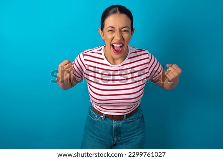 Portrait of beautiful woman wearing striped T-shirt over blue studio background looks with excitement at camera, keeps hands raised over head, notices something unexpected reacts on sudden news.