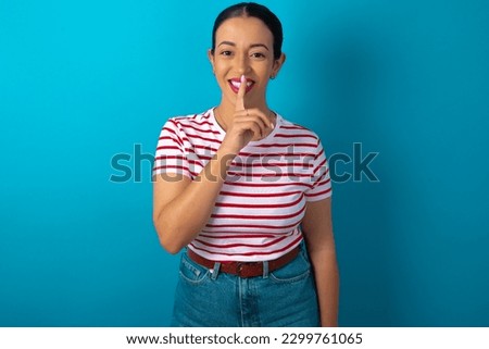 Smiling woman wearing striped T-shirt over blue studio background makes shush gesture, holds fore finger over lips hides secret. Be mute, please.