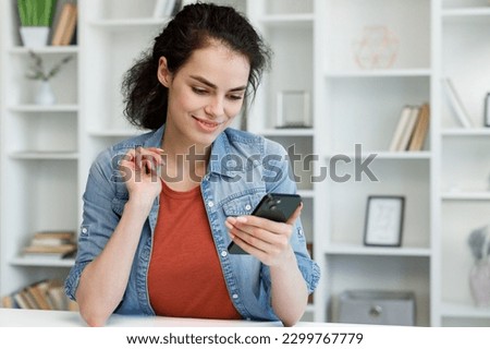 Young curly brunette sitting at the table uses a mobile phone. Surfs the Internet, communicates in instant messengers on his smartphone. Online communication, smartphone in human life. Royalty-Free Stock Photo #2299767779