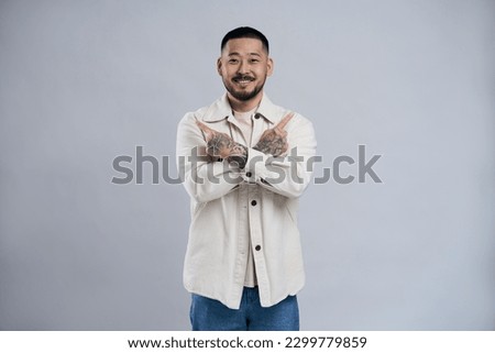 Attractive bearded man pointing up with both fingers, having crossed arms, presenting copy space. Indoor studio shot isolated on grey background