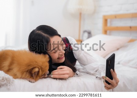 Beautiful asian woman holding her phone up to take a picture with her furry dog on the bed.