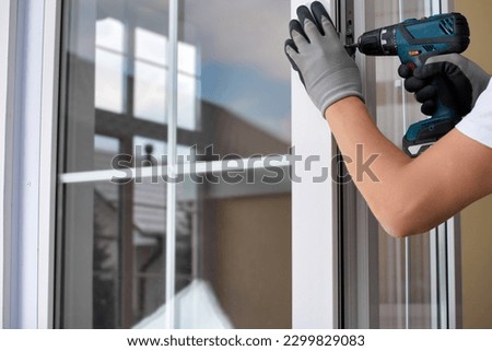 Worker's gloved hand with an automatic screwdriver screws the fastening on the plastic door. An experienced master maintains plastic windows and doors with the help of professional tools Royalty-Free Stock Photo #2299829083