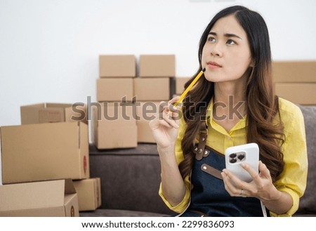 Young asian online seller businesswoman working shipping at home looking at mobile phone for online orders and thinking about her business future, good for online seller business concept. Royalty-Free Stock Photo #2299836093