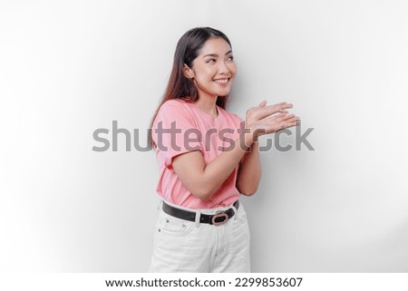 Excited Asian woman is wearing pink t-shirt, pointing at the copy space beside her, isolated by white background Royalty-Free Stock Photo #2299853607