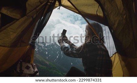 A girl hiker dressed in a plaid shirt sits resting in a yellow tent, takes a selfie on a smartphone. Camping, outdoor adventure lifestyle. Hiker girl taking video selfie, having video chat and smiling