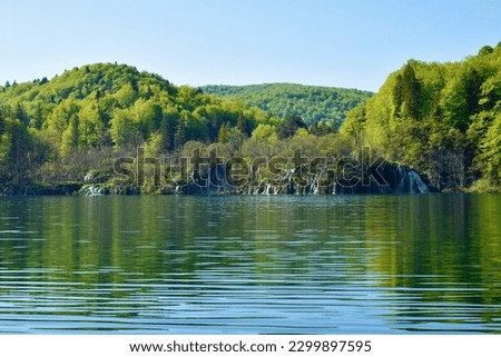 Waterfalls flowing into Kozjak lake at Plitvice lakes in Lika-Senj county, Croatia with forest covered hills above Royalty-Free Stock Photo #2299897595