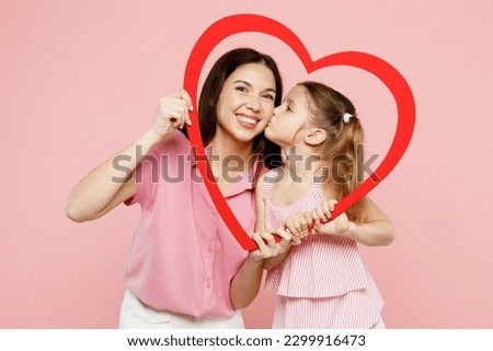 Happy woman wear casual clothes with child kid girl 6-7 years old. Daughter kiss mother hold in hands look through big paper heart frame isolated on plain pink background. Family parent day concept