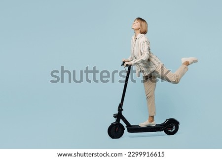 Full body side view healthy happy elderly woman 50s years old wearing shirt riding e-scooter raise up leg isolated on plain pastel light blue cyan color background studio portrait. Lifestyle concept