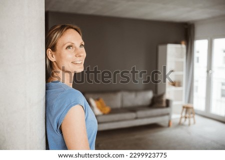 Happy 30-year-old blonde woman in blue t-shirt leaning against a pillar in her apartment and smiling Royalty-Free Stock Photo #2299923775