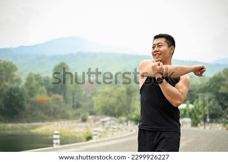 A mature Asian man in sportswear warming up his body before workout, stretching his arms muscle, exercising at a park. Lifestyle concept Royalty-Free Stock Photo #2299926227