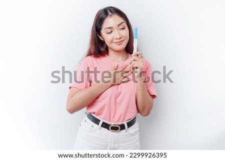 Happy mindful thankful young woman wearing pink t-shirt showing her pregnancy test, hand on chest smiling isolated on white background 