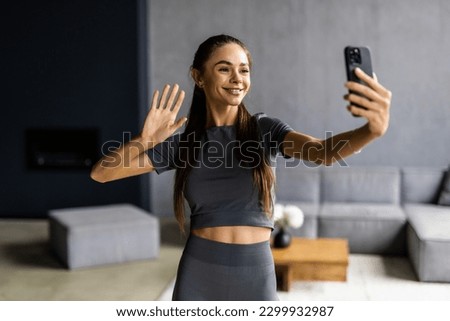 Close up portrait of sportive powerful woman dressed in tight gray top demonstrating her biceps taking selfie or having video call at home