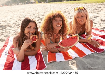 Three beautiful young women lying on the beach sunbathing in swimming suites, eating fruit, relaxing and enjoying.