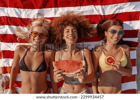 Three beautiful young women lying on the beach sunbathing in swimming suites, eating fruit, relaxing and enjoying. Royalty-Free Stock Photo #2299934457