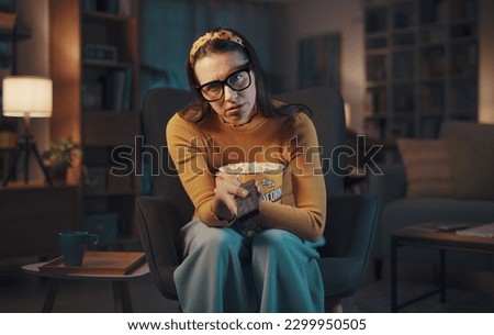 Bored sad woman sitting in the living room and watching TV, she is holding the remote control and looking at camera