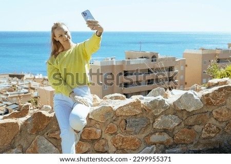 a girl with blond hair holds a phone in her hands and takes a selfie overlooking the sea, the girl is dressed in a yellow jacket and white pants
