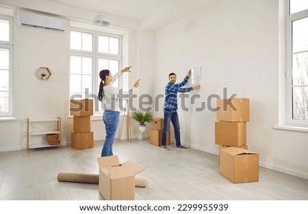 Handsome husband hangs a picture on the wall adjusting the level on the advice of his attractive wife. Young happy married couple takes things out of boxes after moving into new modern apartment.