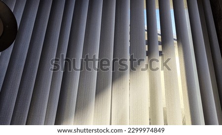 Vertical fabric white blinds on the window with light and shadow from sun. Background and texture. Abstract pattern and frame. Stripes and lines of white-gray color Royalty-Free Stock Photo #2299974849