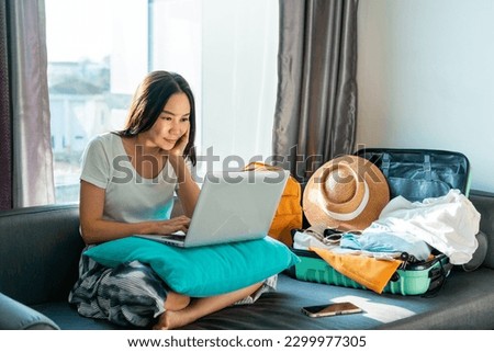 Asian woman prepare things into baggage for vacation trip at home. Happy female using computer to planning travel trip and booking tickets or hotel room on web. Travel, summer and holiday concept. Royalty-Free Stock Photo #2299977305
