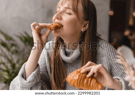 Young blonde woman with bang eating croissants at a cafe. Girl bite piece of croissant look joyful at restaurant. Cheat meal day concept. Woman is preparing with appetite to eat croissant. Royalty-Free Stock Photo #2299982201