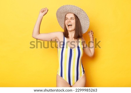 Extremely happy woman wearing striped swimming suit and sun hat isolated on yellow background dancing celebrating her vacation, enjoying sunbathing and swimming in sea.