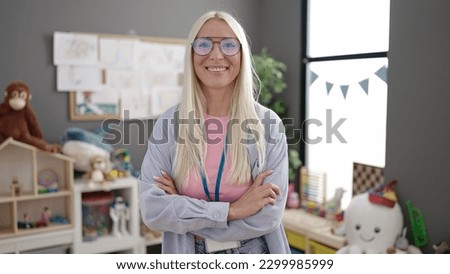 Young blonde woman preschool teacher smiling confident standing with arms crossed gesture at kindergarten
