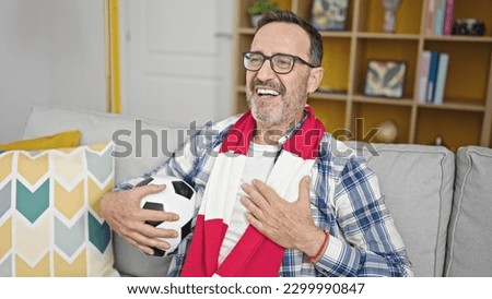 Middle age man supporting soccer team singing hymn with hand on heart at home Royalty-Free Stock Photo #2299990847