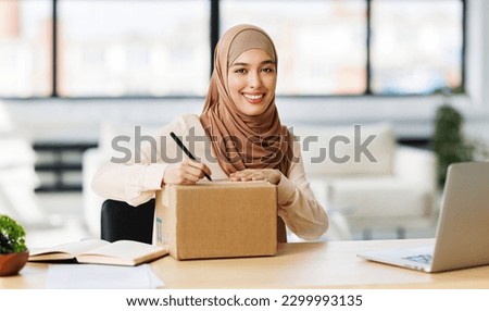 Beautiful young smiling muslim woman in traditional religious hijab   signs carton box to send parcel while at table in home office
