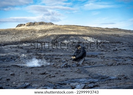 man taking a picture with a phone of a drone on lava field at icelandic Fagradalsfjall volcano with steam rising from under the lava