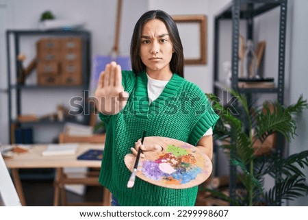 Young south asian woman holding painter palette with open hand doing stop sign with serious and confident expression, defense gesture 