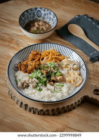 Bubur ayam or chicken porridge is Chinese Indonesian rice porridge topped with various savoury condiments served on white bowl and wooden table. Royalty-Free Stock Photo #2299998849