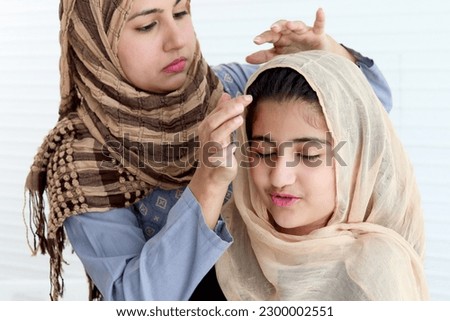 Muslim mother dressing up her daughter girl with traditional hijab scarf, adorable happy smiling kid with beautiful eyes smiling at mom, happy lovely Islamic family on white background.
