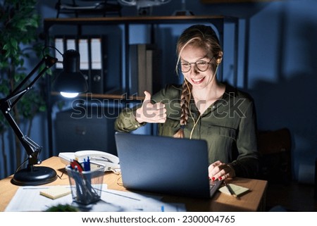 Young blonde woman working at the office at night doing happy thumbs up gesture with hand. approving expression looking at the camera showing success. 