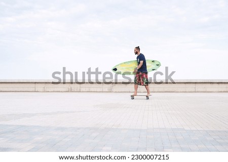 Side view distant guy standing barefoot on longboard riding on paved embankment while holding surfing board in arms against cloudy sky