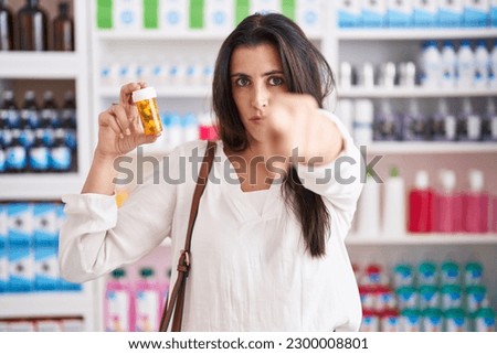 Young brunette woman shopping at pharmacy drugstore holding pills pointing with finger to the camera and to you, confident gesture looking serious 