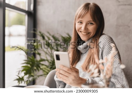 Young blonde woman with bang using phone sits in cafe at table hold smartphone, answering texts, phone call, letters, posting photos, chatting or reading sms. Girl messaging, paying using phone. Royalty-Free Stock Photo #2300009305