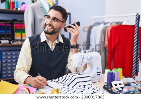 Young arab man tailor listening voice message by smartphone writing on notebook at clothing factory