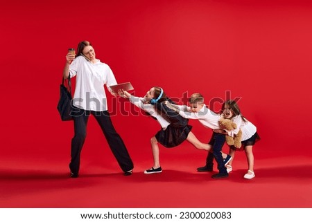 Portrait of businesswoman, mother talking on phone, little boy and girls, children pulling her hand, disturbing her against red studio background. Concept of family, motherhood, childhood, lifestyle Royalty-Free Stock Photo #2300020083