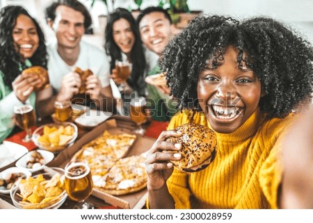 Multiracial happy friends eating hamburger and pizza sitting at pub restaurant table - Young people having diner party in house balcony - Delivery food concept with guys and girls dining in apartment 