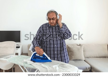 Senior man listening to music while ironing at home. Senior man with headphones ironing. 