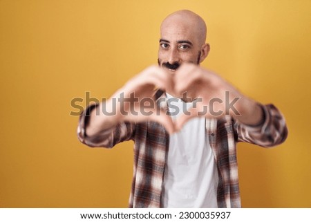 Hispanic man with beard standing over yellow background smiling in love doing heart symbol shape with hands. romantic concept. 
