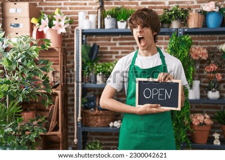 Hispanic young man working at florist holding open sign angry and mad screaming frustrated and furious, shouting with anger. rage and aggressive concept. 