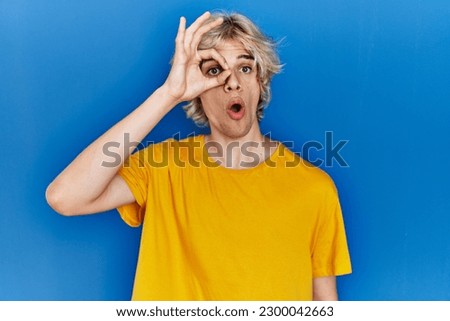 Young modern man standing over blue background doing ok gesture shocked with surprised face, eye looking through fingers. unbelieving expression. 