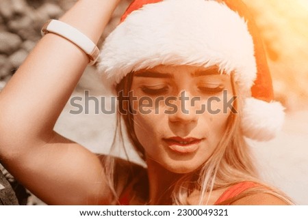 Woman summer travel sea. Happy tourist in red bikini and Santas hat enjoy taking picture outdoors for memories. Woman traveler posing on the beach surrounded by volcanic mountains, sharing travel joy
