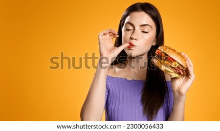 Brunette girl licks her finger, eats tasty hamburger. Woman orders takeaway food with delivery app, enjoys delicious burger, orange background. Restaurants and takeout concept Royalty-Free Stock Photo #2300056433