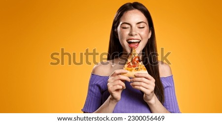 Excited 25 years woman bites tasty slice of pizza, orders delivery takeout from restaurant, hungry girl eats isolated on orange background. Royalty-Free Stock Photo #2300056469