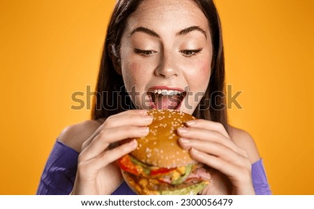 Close up of beautiful woman bites big delicious burger. Hungry girl eats hamburger with pleasure, looks with appetite at junk food, breaking diet, stands over orange background.