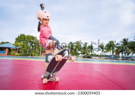 asian child skater or kid girl playing skateboard or ride surf skate and ollie jumping fun or raise wheel manual in skate park by extreme sports to wears helmet elbow pads wrist knee guard body safety Royalty-Free Stock Photo #2300059403